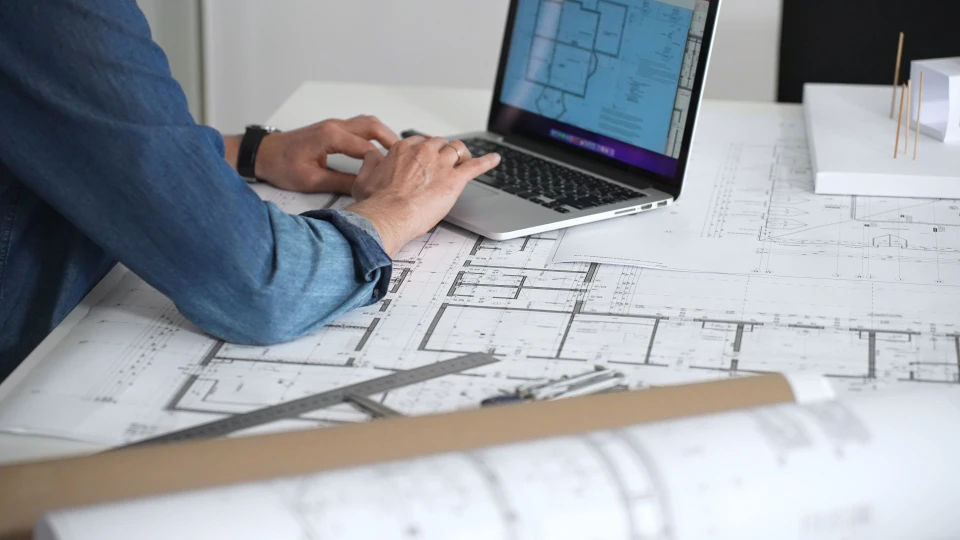 Intérieur bureau lumineux avec fenêtre, personne en chemise vichy travaillant sur plans architecturaux étalés sur table